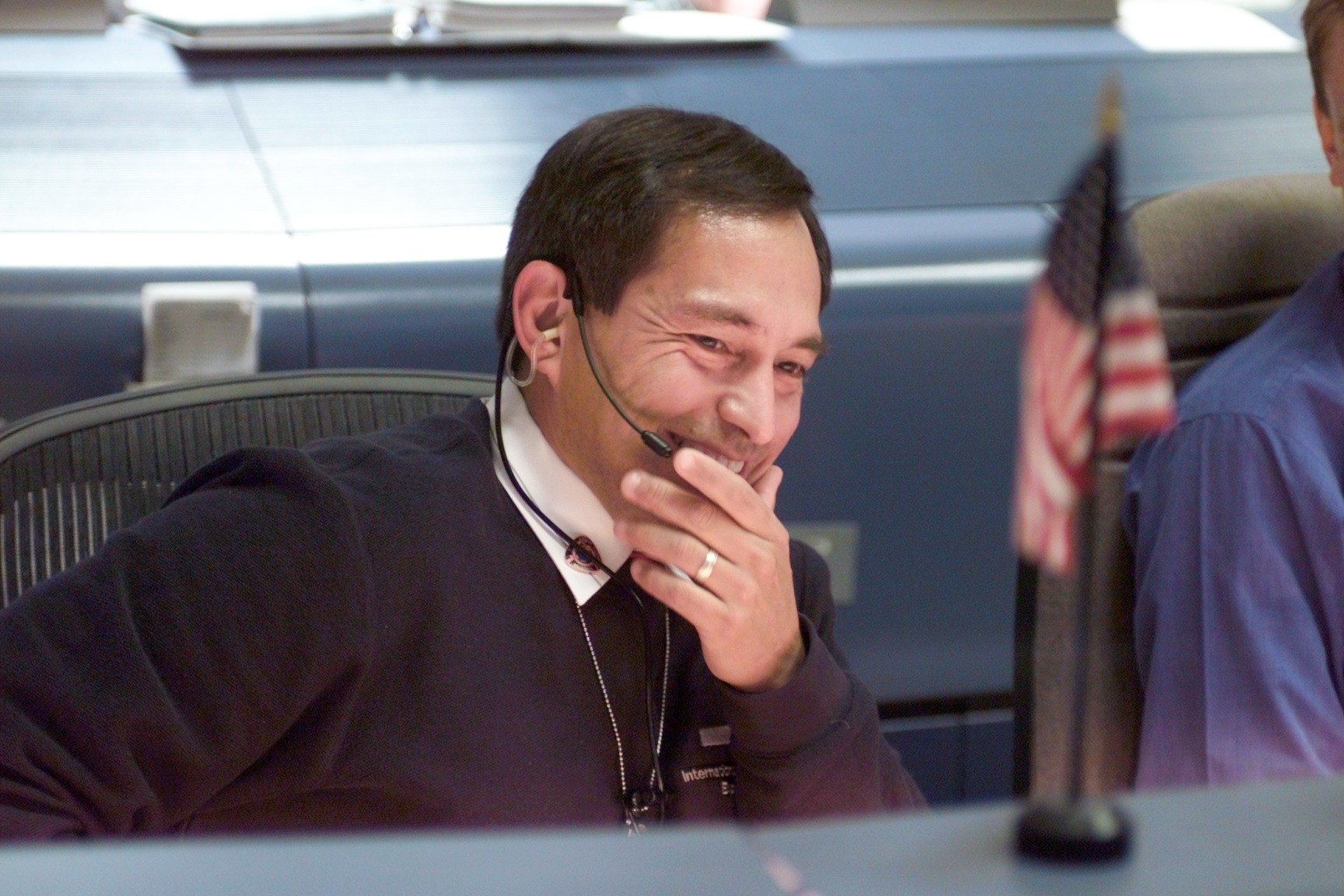 A man with dark hair sits in a chair, his hand partially covering his smile. He wears a black sweater with some words embroidered on it, a white shirt with a pin on the collar, a wedding ring, and a headset with a microphone. In the foreground is a small American flag on top of a desk.