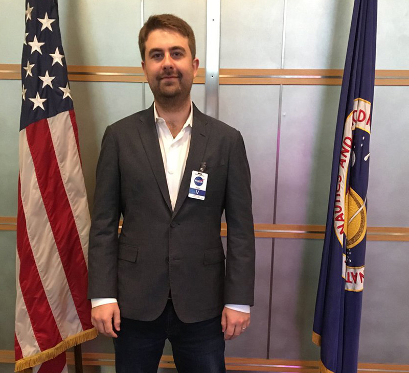 A young man in a suit is standing between the American flag and a NASA flag.