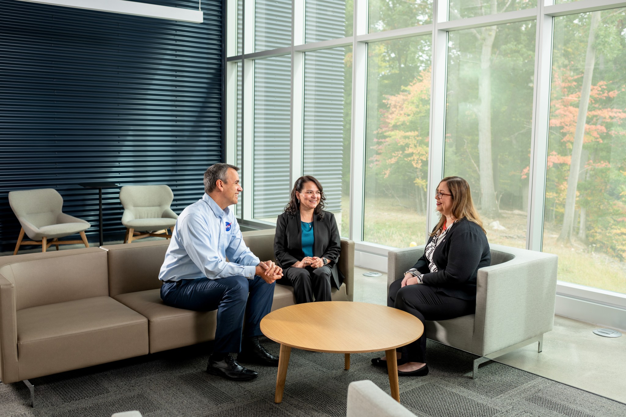 Nelson Morales (left), Janette C. Briones (center), and Azlin Biaggi-Labiosa smile at one another and sit on couches around a circular wooden table inside NASA Glenn Research Center’s Aerospace Communications Facility. Autumn leaves can be seen outside a large glass window in the background.