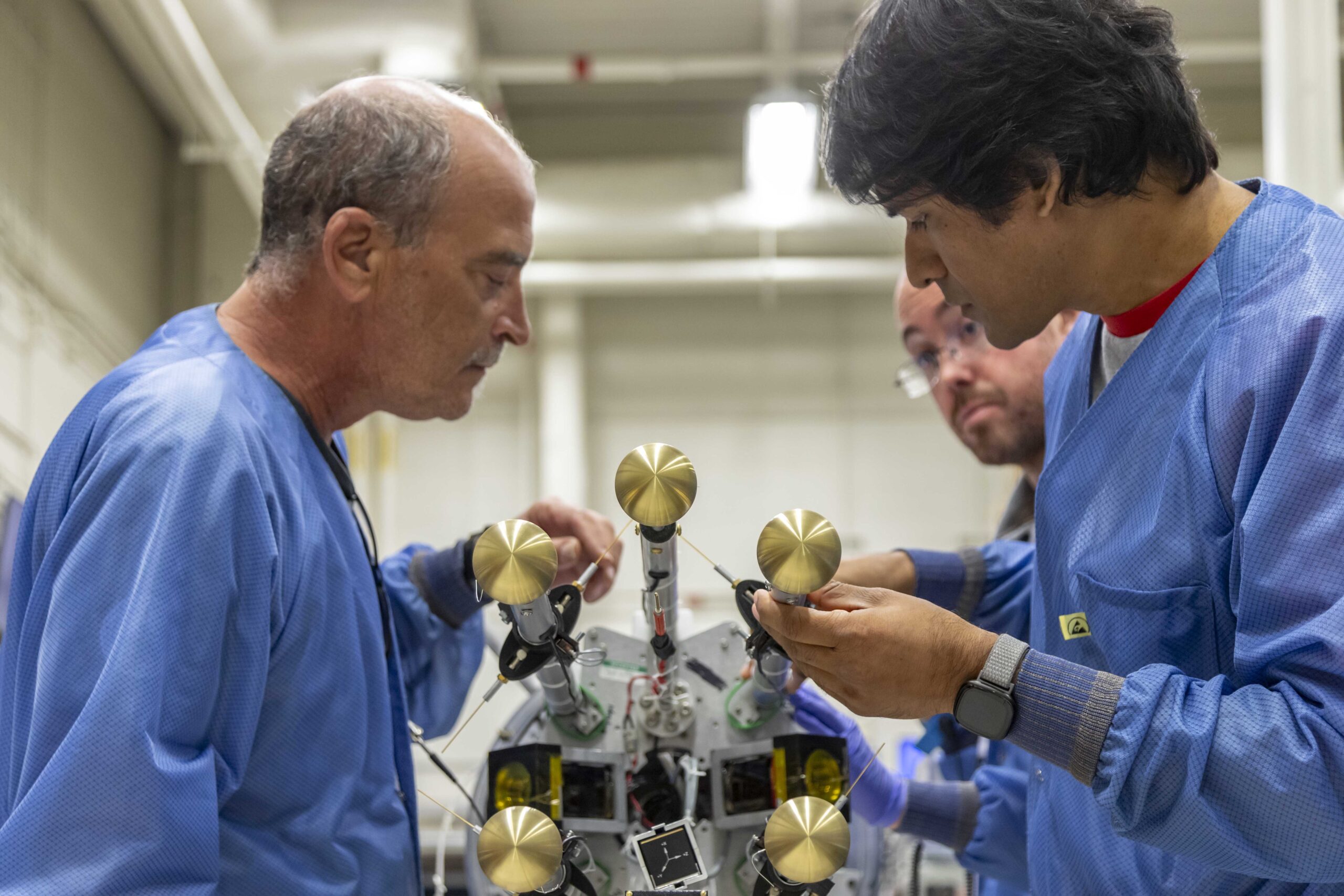 Three men stand over a rocket laid down on a table in front of them. Two in the foreground are adjusting a gold-colored metal rod protruding from the end of the rocket.