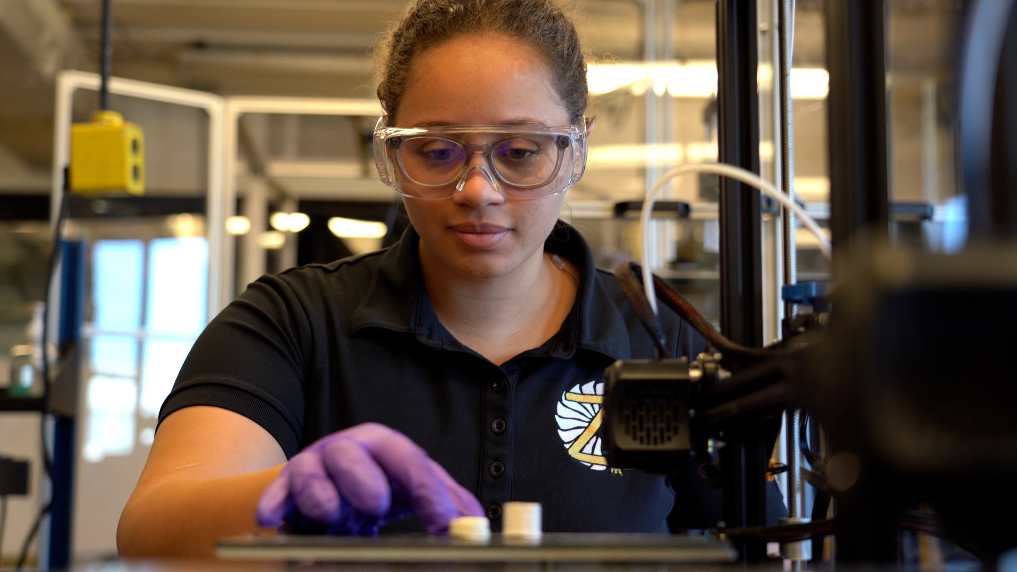 A female scientist stands in a research laboratory wearing glasses and safety goggles. Her right-hand, covered by a purple glove, hovers over a black table as she gently touches and inspects two small 3D printed samples that are white. One sample is shorter than the other.